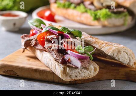 Beef baguette sandwich with tomatoes, basil, red onion served on ceramic plate with ingredients above over grey blue table. Stock Photo