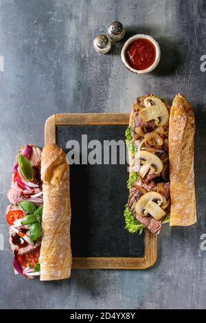 Variety of Beef baguette sandwiches with champignon mushrooms, green salad, onion, tomatoes served on empty black chalkboard with knife, salt and sauc Stock Photo