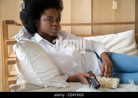 Focused African American biracial millennial woman lying in bed, relaxing, eating popcorn while watching movie on tv, holding remote controller. Lazy Stock Photo