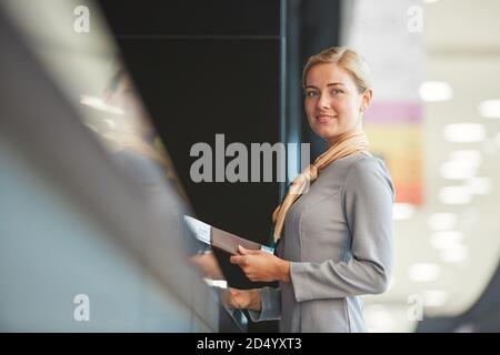 Waist up portrait of elegant flight attendant standing by check in desk and smiling at camera holding tickets, copy space Stock Photo