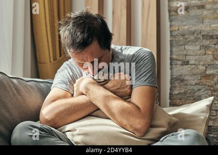 Depressed man sitting on living room sofa and hugging pillow, portrait of adult male having anxiety attack Stock Photo