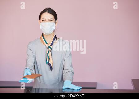 Waist up portrait of smiling flight attendant wearing mask while standing at check in desk handing tickets to passenger, copy space Stock Photo