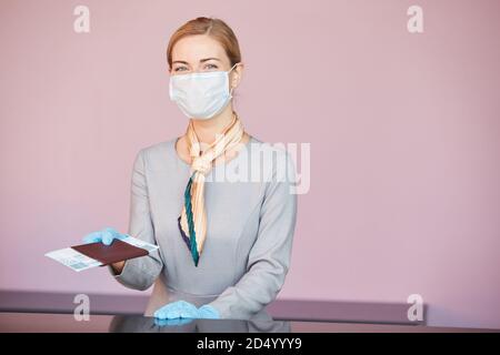 Waist up portrait of blonde flight attendant wearing mask while standing at check in desk handing tickets to passenger, copy space Stock Photo