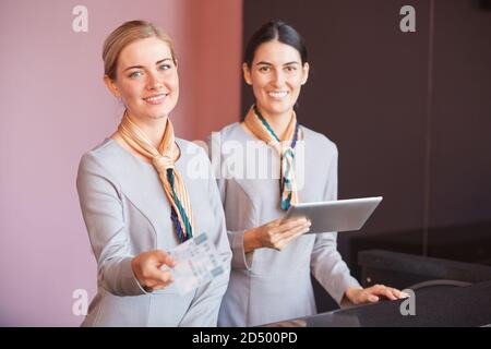Portrait of two smiling flight attendants handing tickets to passenger while standing at check in desk in airport, copy space Stock Photo