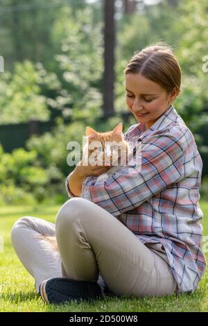 Close up of smiling woman in checked shirt looking at camera, hugging and embracing with tenderness and love domestic ginger cat outdoors. Love to the Stock Photo