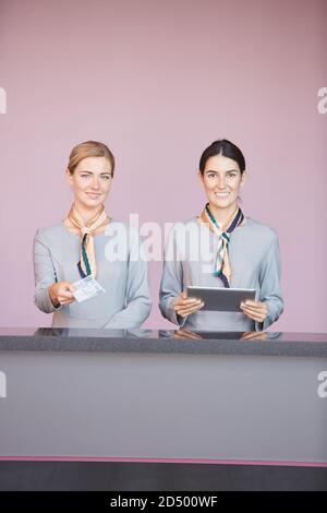 Vertical portrait of two smiling flight attendants handing tickets to passenger while standing at check in desk in airport, copy space Stock Photo