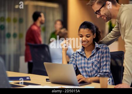 Young cheerful caucasian man helping female coworker with computer work, two young multiracial colleagues using laptop and discussing project. Teamwork, cooperation, business concept Stock Photo