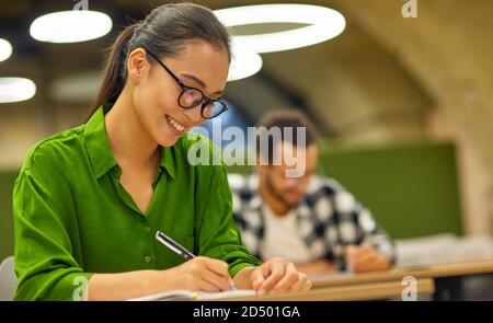 Young happy asian woman, female office worker sitting at desk in the office or coworking space and making some notes during corporate seminar or training. Business people and education Stock Photo