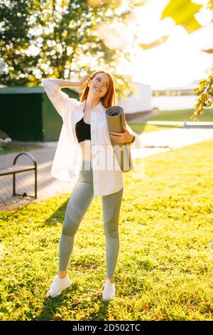 Portrait of cheerful attractive red-haired young woman with yoga mat, standing at city park. Stock Photo