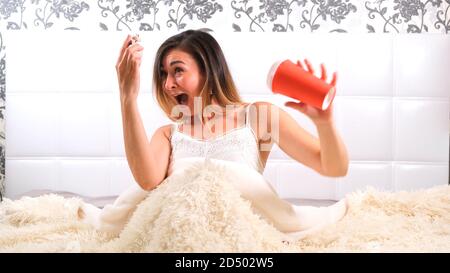 Attractive young woman gets up in bed with a cup of coffee and an alarm clock. The woman overslept and is late, reacting with horror at the time. Stock Photo