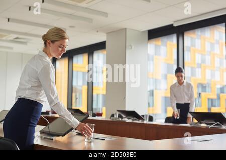 Portrait of female assistant putting glass of water on table while preparing conference room for business event, copy space Stock Photo