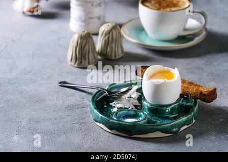 Breakfast with cup of coffee and soft boiled egg, served in green ceramic egg cup with salt, pepper and toasted bread, jug of cream over grey blue tab Stock Photo