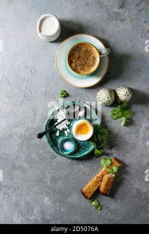 Breakfast with cup of coffee and soft boiled egg, served in green ceramic egg cup with salt, pepper and toasted bread, jug of cream over grey texture Stock Photo