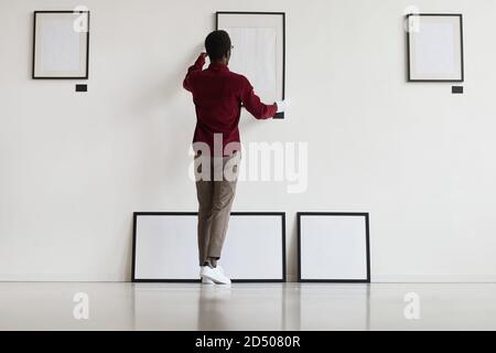 Full length back view at African-American man hanging blank frames on wall while planning art gallery or exhibition, copy space Stock Photo