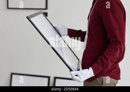 Cropped portrait of unrecognizable man wearing gloves holding painting frame while standing against white wall in art gallery, copy space Stock Photo