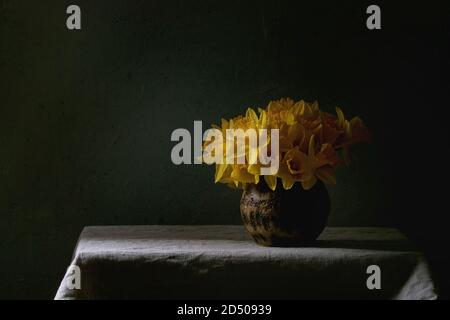 Narcissus daffodils flowers in ceramic vase standing on table with linen tablecloth. Dark still life. Copy space Stock Photo