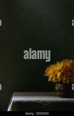 Narcissus daffodils flowers in ceramic vase standing on table with linen tablecloth. Dark still life. Copy space Stock Photo