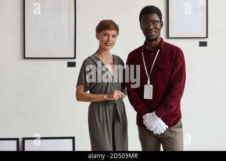 Portrait of female art gallery manager posing with African-American worker while standing against white wall and smiling at camera, copy space Stock Photo