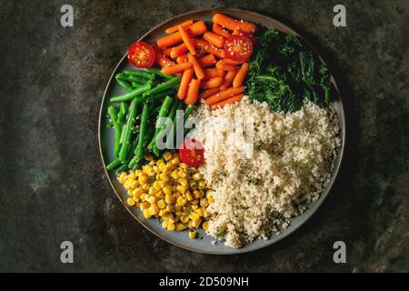 Couscous with parboiled vegetables baby carrots, green beans, sweet corn, spinach served in ceramic plate with cherry tomatoes and sesame seeds. Vegan Stock Photo