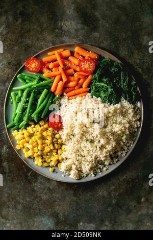 Couscous with parboiled vegetables baby carrots, green beans, sweet corn, spinach served in ceramic plate with cherry tomatoes and sesame seeds. Vegan Stock Photo