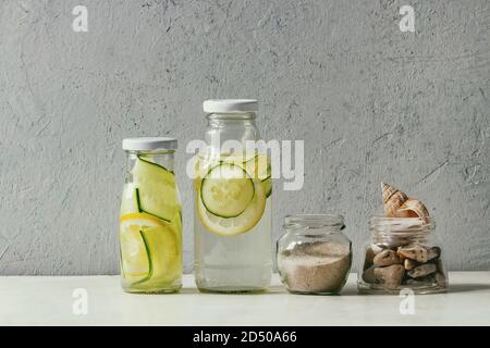 Summer theme. Two glass bottles with lemon and cucumber infusion sassy water, seashells and sand in glass jars on white marble table, concrete wall at Stock Photo
