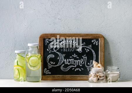Summer theme. Chalk lettering Hello summer on vintage chalkboard, seashells and sand in glass jars, sunglasses, two glass bottles with sassy water on Stock Photo
