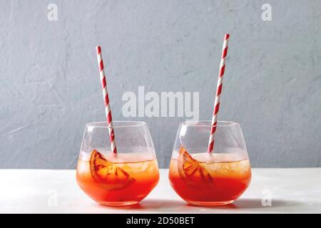 Blood orange iced cocktails in glasses, decorated by slice of oranges, served with retro straw on white marble table with grey wall at background. Stock Photo