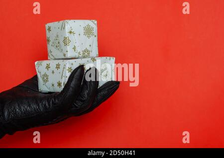 A hand in a black leather glove holds a wrapped Christmas present on a red background. Small rectangular box wrapped in white wrapping paper. Christma Stock Photo