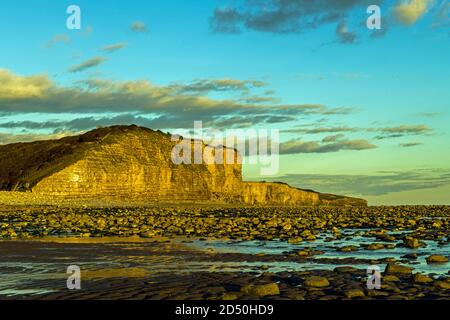 The Beach at Llantwit Major, also known as Colhuw or Colhugh Beach. Behind the cliffs glow in the late evening sun behind the sand and rocks. Stock Photo