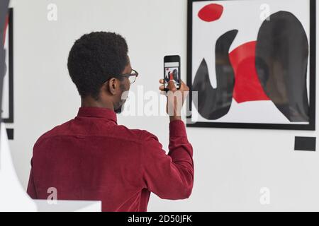 Back view portrait of young African-American man taking photo of painting via smartphone at contemporary art gallery exhibition, copy space Stock Photo