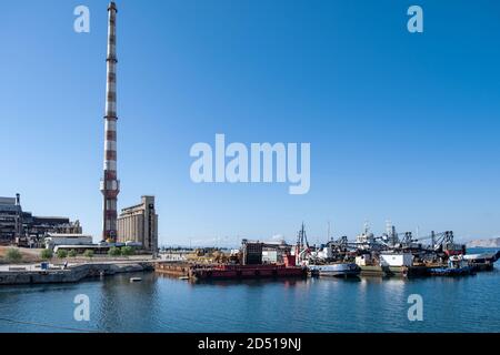 Old abandoned factory industrial plant and boats anchored in harbor. Fertilizer factory ruins in Drapetsona Piraeus Greece, blue sky and sea, sunny da Stock Photo