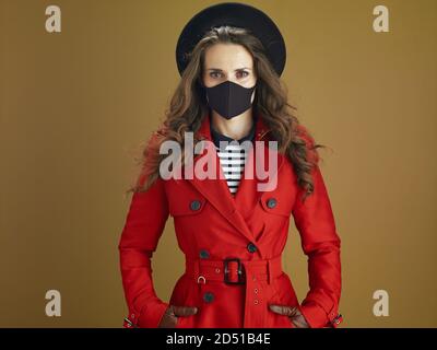 Hello autumn. Portrait of trendy middle aged housewife in red coat with black mask isolated on beige. Stock Photo
