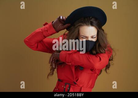 Hello autumn. modern 40 years old woman in red coat with black mask sneezing into elbow on bronze background. Stock Photo