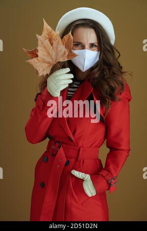 Life during covid-19 pandemic. happy young woman in red coat with yellow autumn maple leaf isolated on beige background. Stock Photo