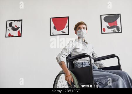 Wide angle portrait of young woman using wheelchair and wearing mask while looking at paintings in modern art gallery Stock Photo