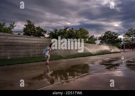 young girl jumps in sprinkler puddles of water on the sidewalk Stock Photo