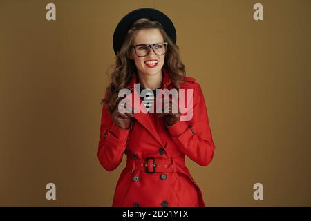 Hello november. smiling modern 40 years old woman in red coat and black beret with leather gloves and glasses looking into the distance on brown backg Stock Photo