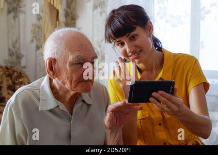 A grandfather and an adult granddaughter are sitting on the sofa and looking at their mobile phone. A young woman teaches an older man to use a modern Stock Photo