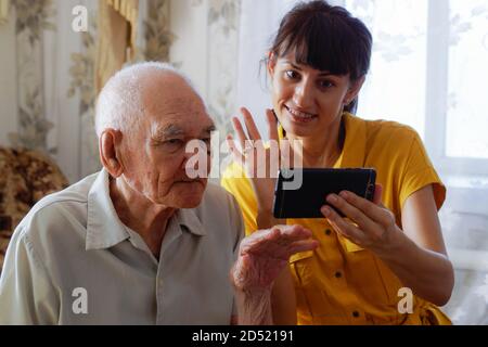 An elderly grandfather and a young woman have fun using smartphone apps granddaughter teaches grandfather online entertainment, online communication. Stock Photo