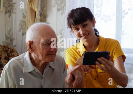 A grandfather and an adult granddaughter are sitting on the sofa and looking at their mobile phone. A young woman teaches an older man to use a modern Stock Photo