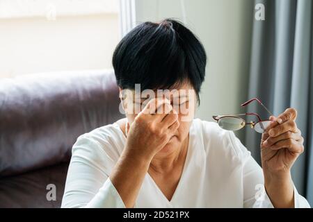 Tired old woman removing eyeglasses, massaging eyes after reading paper book. feeling discomfort because of long wearing glasses, suffering from eyes Stock Photo