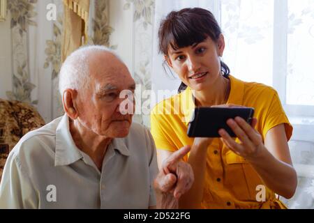 a man of the 90s with an adult granddaughter, sitting together on a cozy sofa with a phone in his hands, looking at photos. a woman in a yellow dress Stock Photo