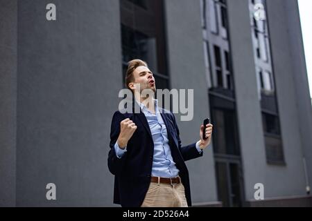 Excited young man celebrating victory. Big success, great achievements, positive emotions. Handsome businessman with smartphone getting good news Stock Photo