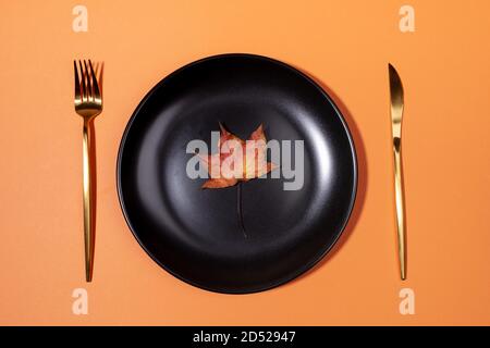 Top view one fall dried colorful maple leaf on black plate with golden fork and knife on trendy pastel orange backdrop. Stock Photo