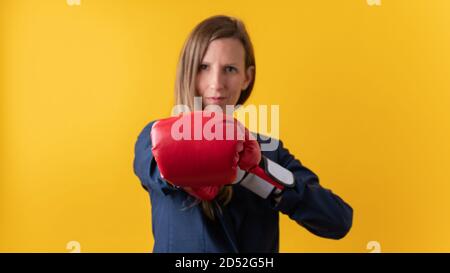 Confident young businesswoman wearing red boxing gloves punching towards the camera. Over yellow background. Stock Photo