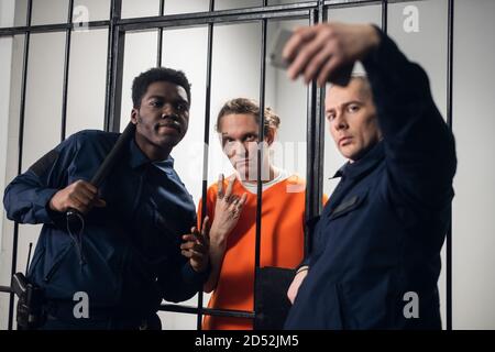 Wardens in a maximum security prison take selfies with a prisoner with tattoos on his face, who is sitting in a cell Stock Photo