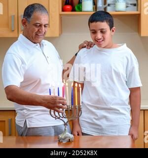 Happy family is lighting a candle celebrating together Jewish holiday Hanukkah. Jewish Dad and teenager son or grandfather with grandson lighting Chanukkah Candles in a menorah for the holdiay Stock Photo