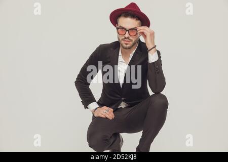 Tough fashion model fixing his sunglasses and looking away, wearing hat while crouching on gray studio background Stock Photo