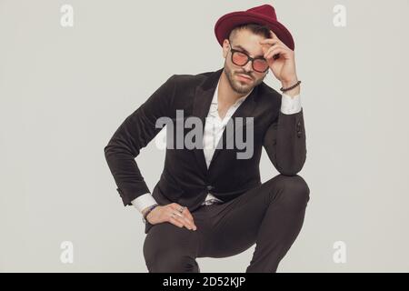 Tough fashion model fixing his hat and wearing sunglasses while crouching on gray studio background Stock Photo