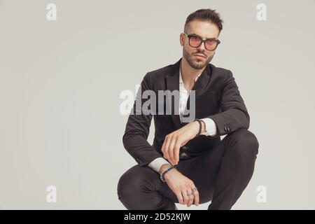 Confident fashion model looking forward and wearing sunglasses while crouching on gray studio background Stock Photo